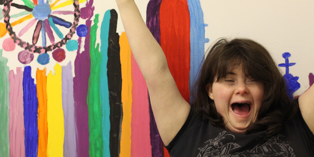 Young woman with Downs syndrome in front of a colourful painting with arms raised in celebration