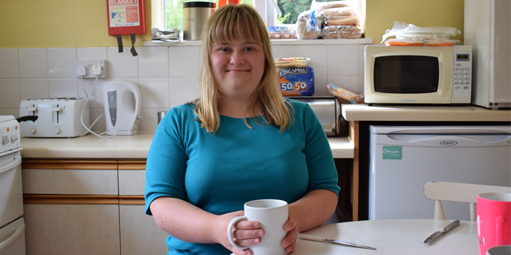 Hanna tea Young woman with Down Syndrome sitting at a kitchen table holding a mug, smiling at the camera