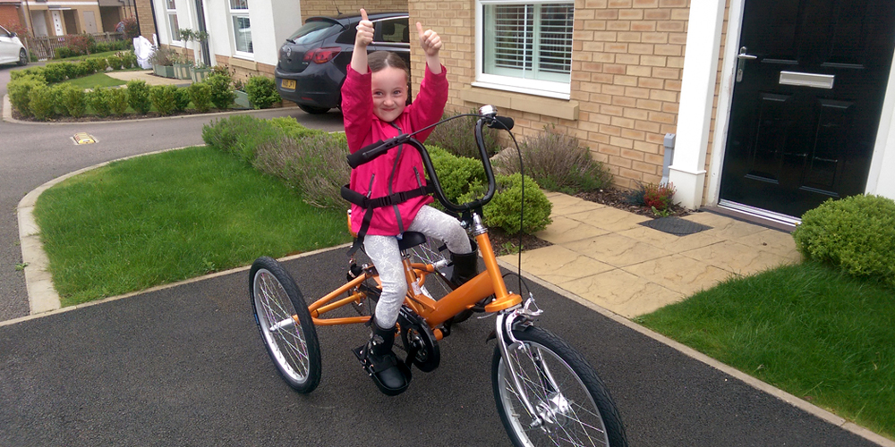 Young girl on an orange adapted trike giving two thumbs up