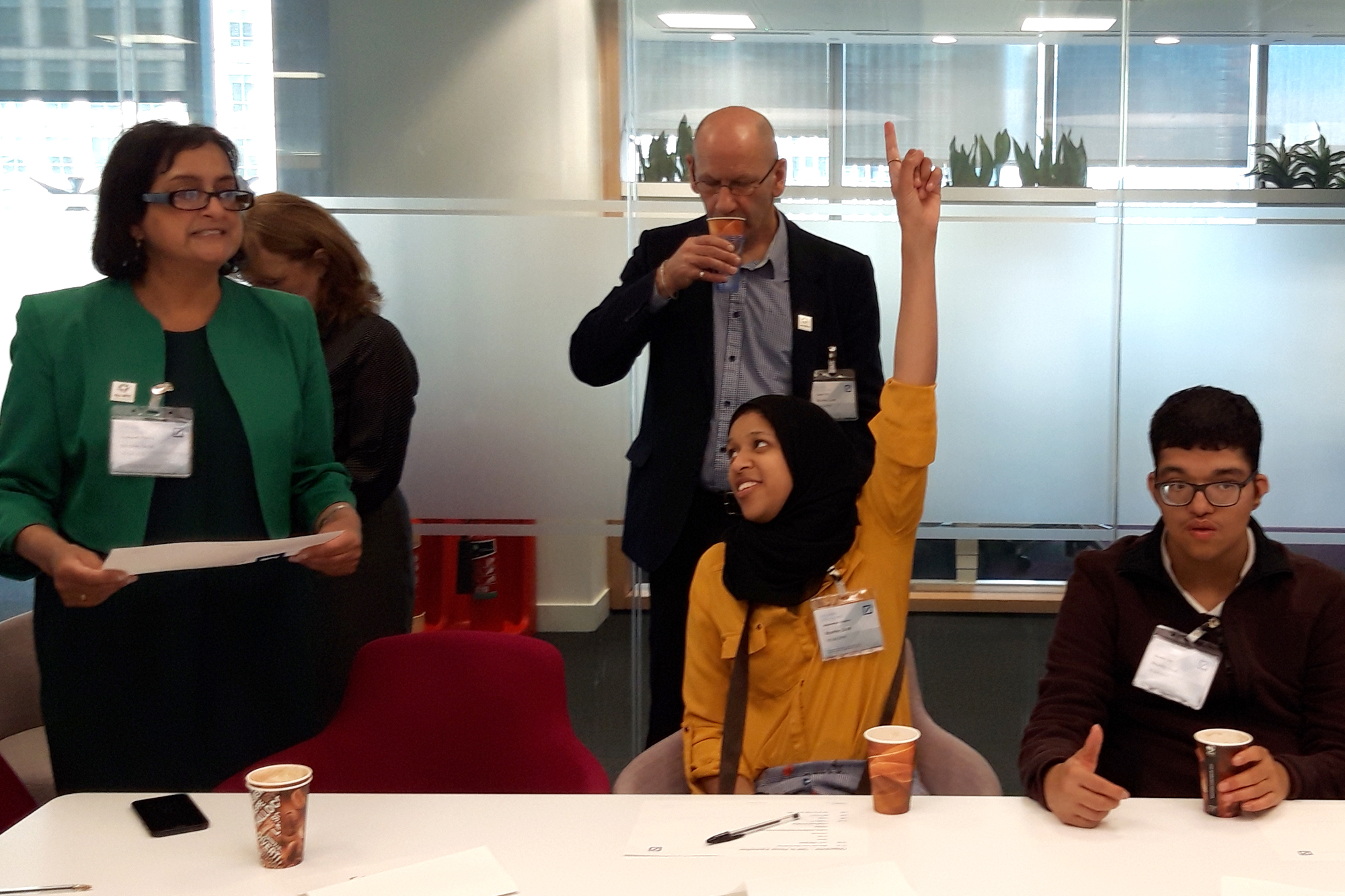 Young woman raising her hand at a conference table with others sitting and standing around her