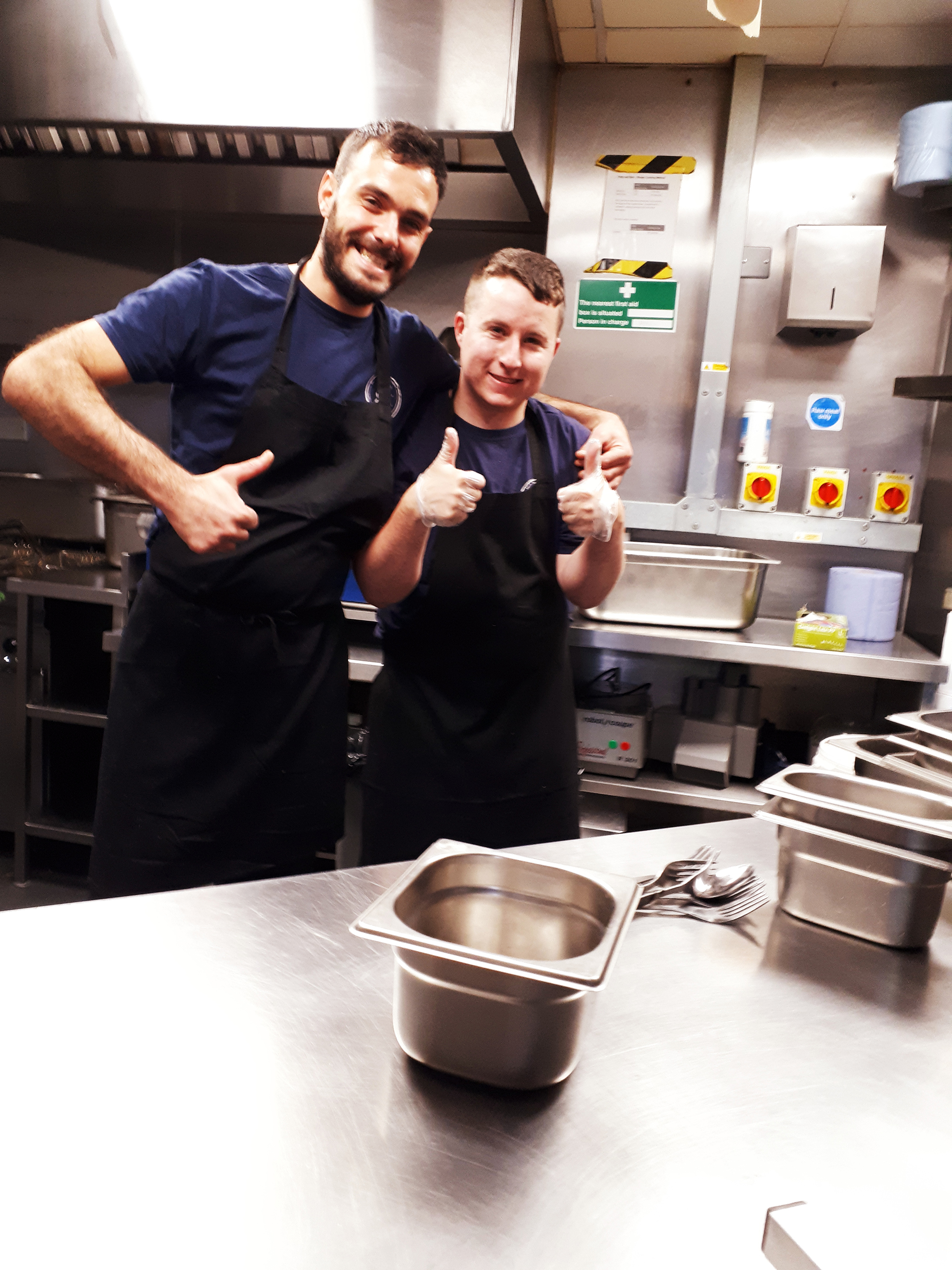Young man and his coworker in a kitchen giving thumbs up
