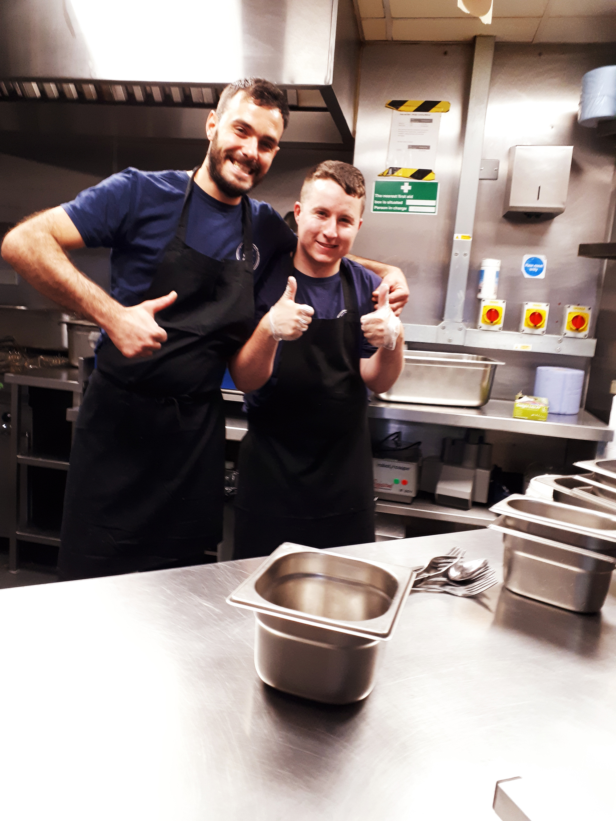 AlexwithhisbuddyMateo Young man and his coworker in a kitchen giving thumbs up