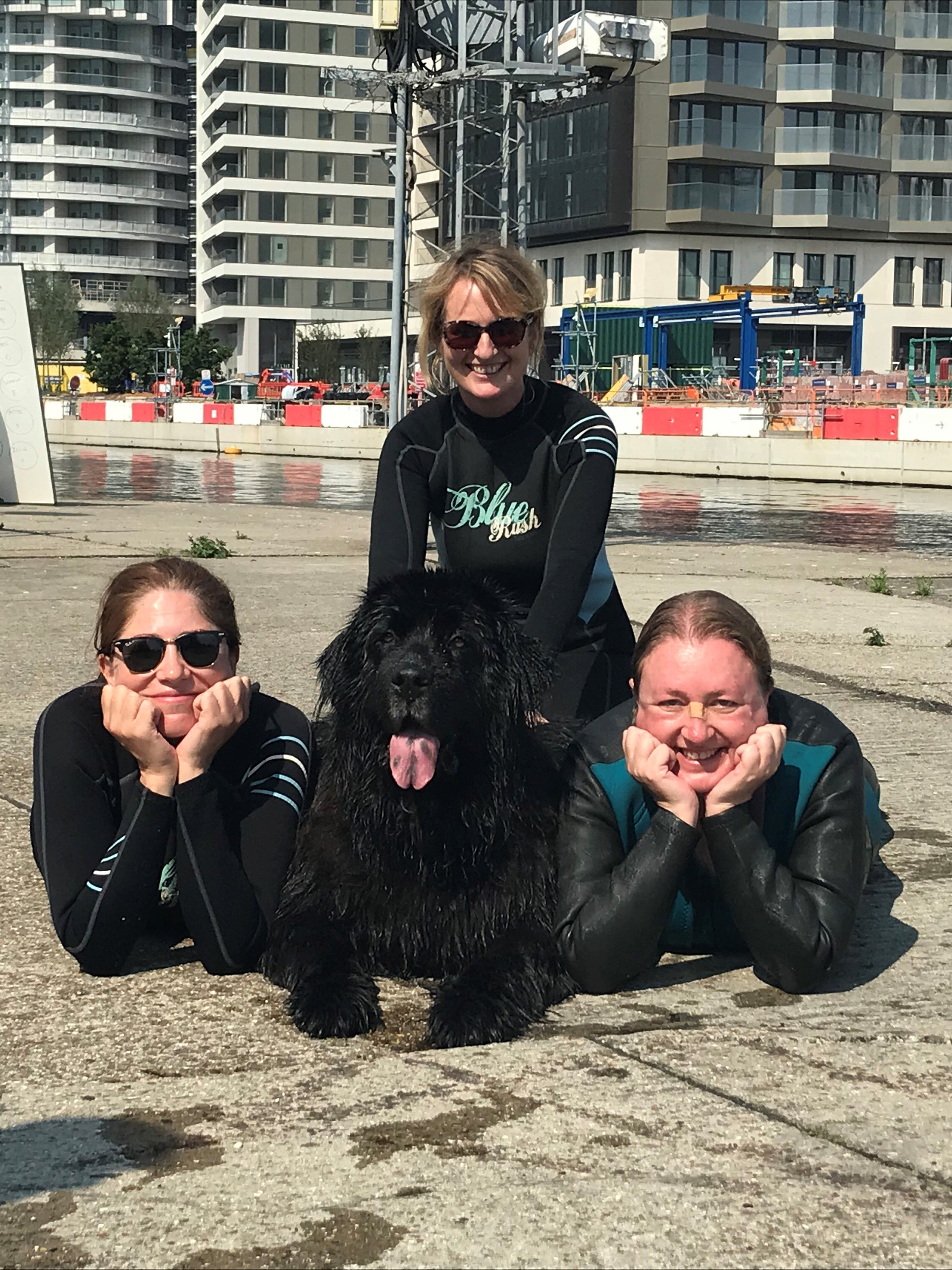 two women are laying down with their head on their hands while another is kneeling behind them, they have a large dog in the middle of them