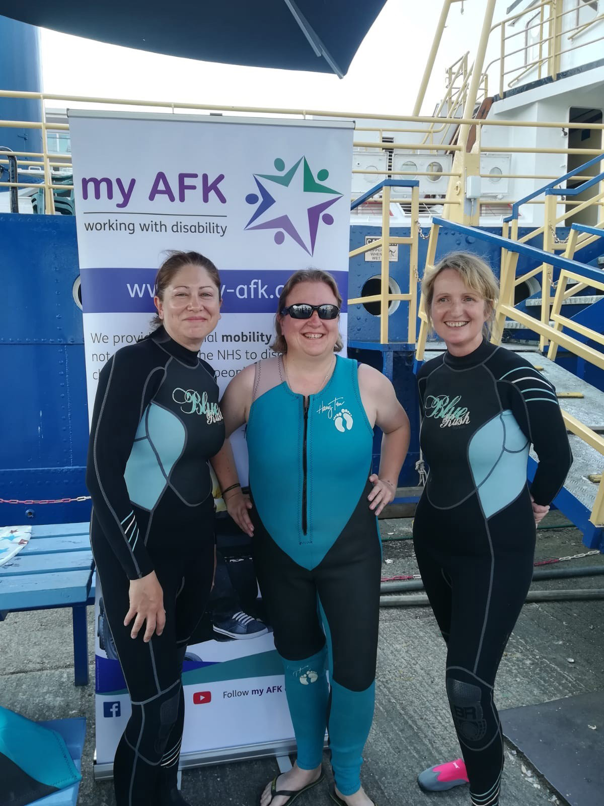 Three women are stood wearing wetsuits in front of a myafk banner