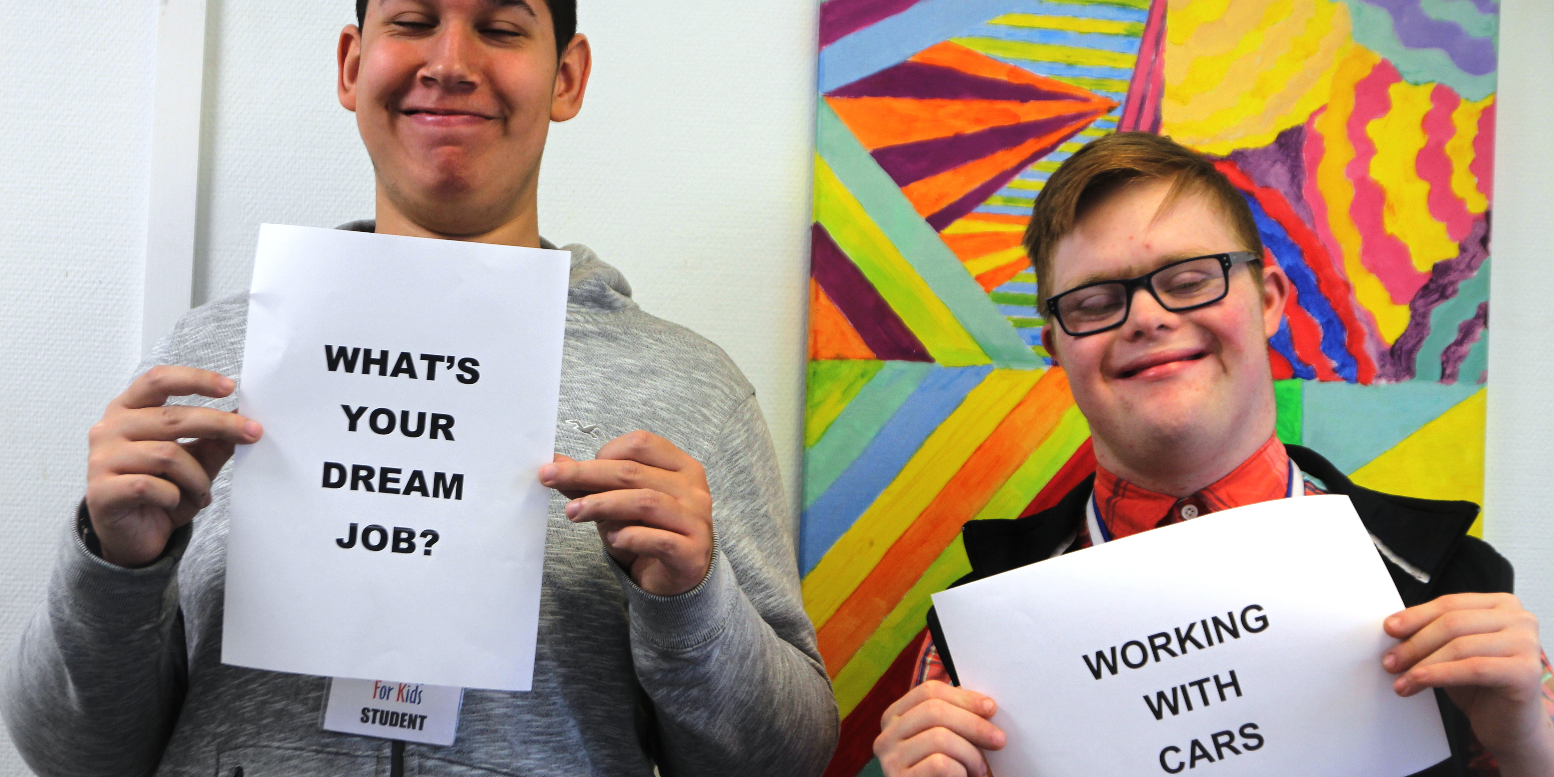 two people are stood, smiling and holding up signs that explain their work experience. One says "What's your dream job" the other says "Working with Cars"