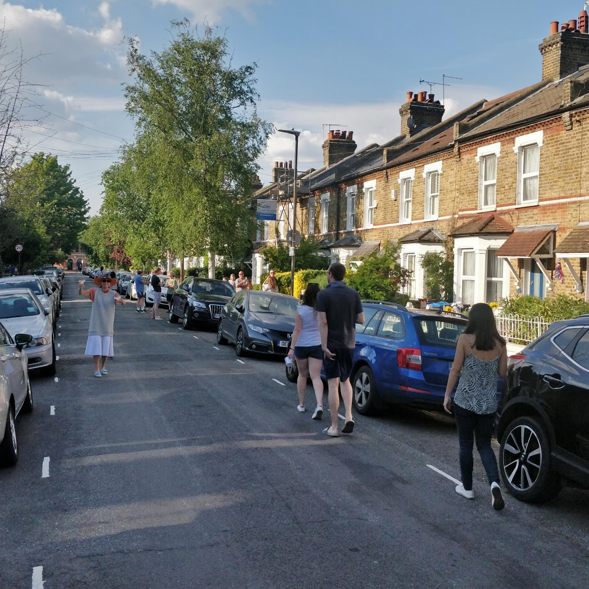 A quiet residential street lined with parked cars, with people walking down the street celebrating VE Day