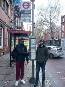 two young men smiling next to a bus stop. One of them is pointing to the timetable.