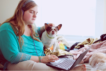 girl sits on her bed with her laptop wearing headphones with a small dog sat next to her.