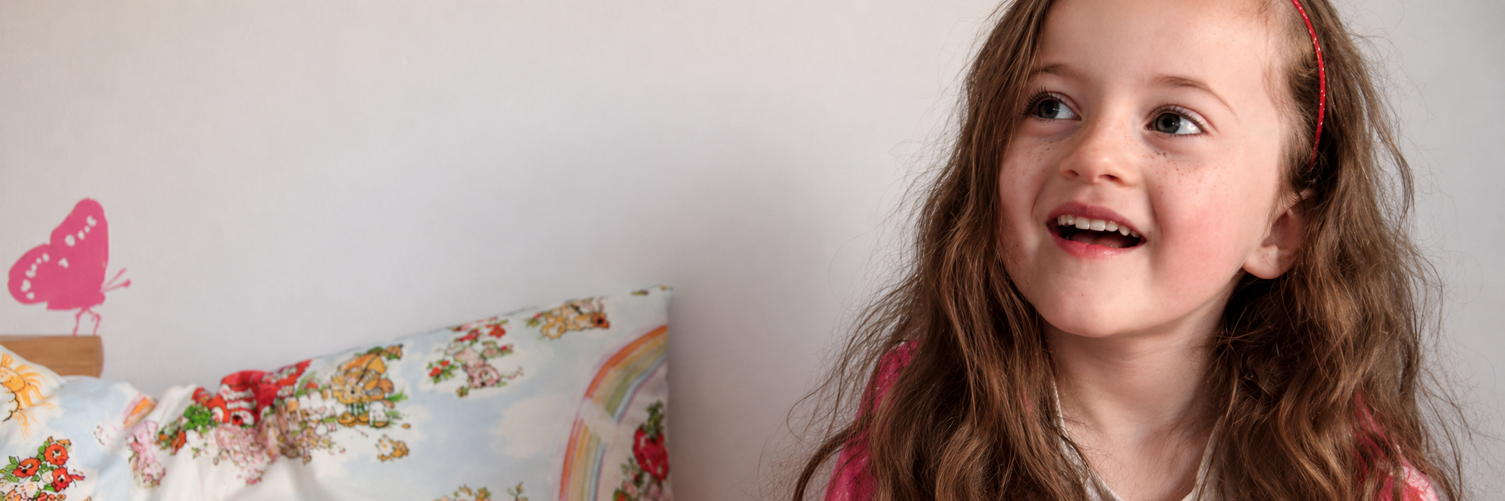 A smiling young girl with long brown hair sits beside a bed with colourful patterned bedding, with a pink butterfly decoration on the wall behind her.