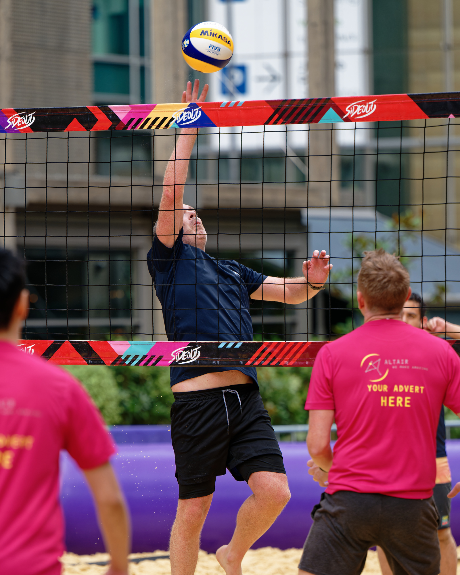 AFK Beach volleyball match action A player reaches up to return a beach volleyball over the net during an AFK Beach match, with teammates in pink shirts watching.