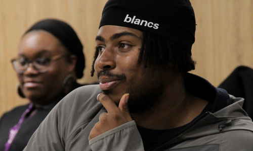 A participant wearing a black beanie listens attentively during a group session, with another attendee blurred in the background.