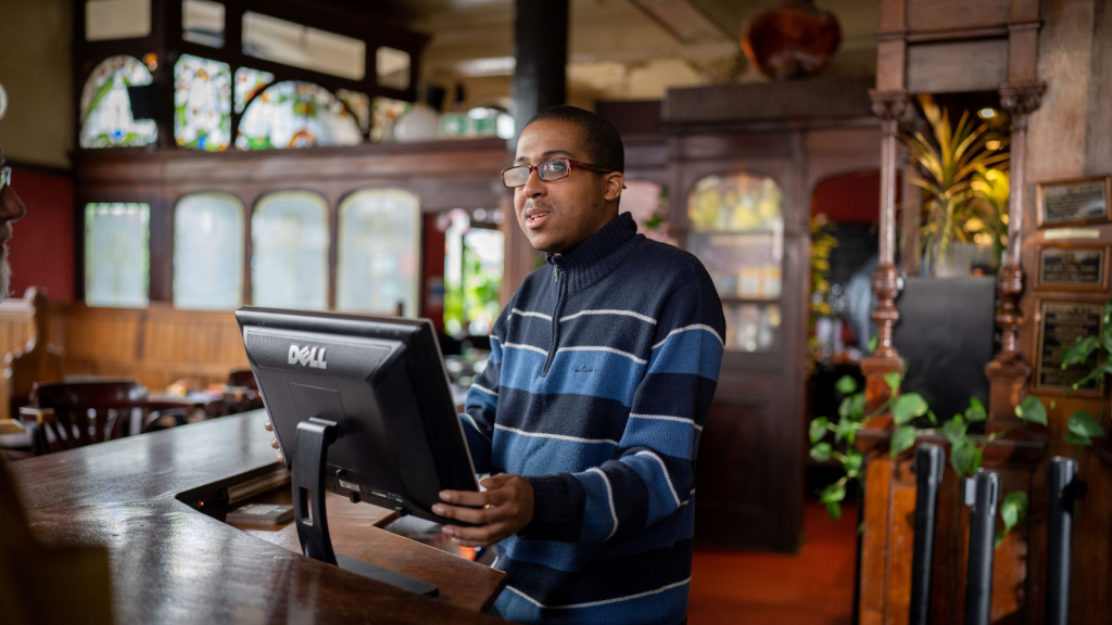 Young man wearing glasses using a touchscreen till behind a wooden bar.